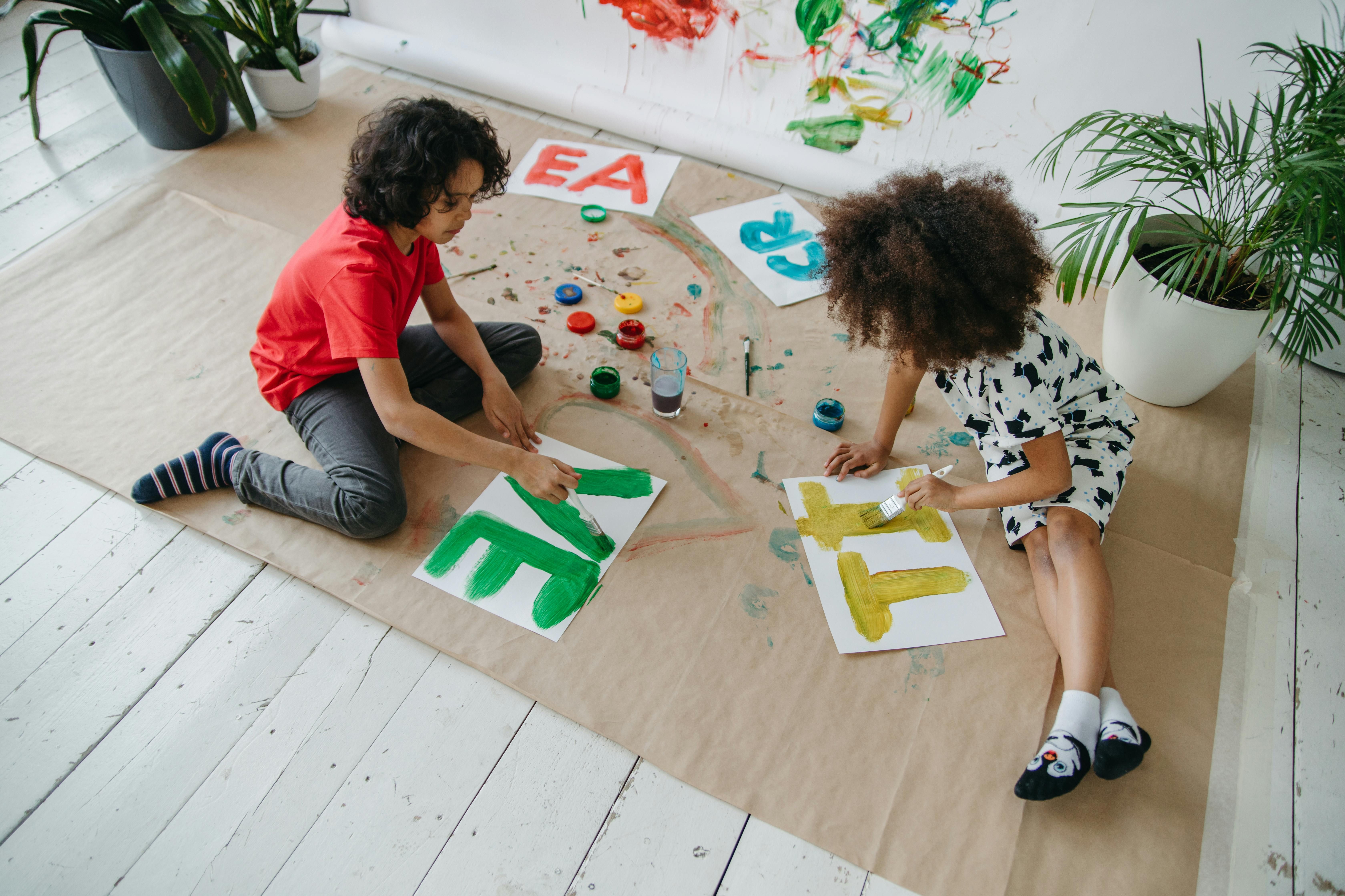 Zwei Kinder sitzen auf dem Boden und malen mit bunten Farben Buchstaben auf weißes Papier.