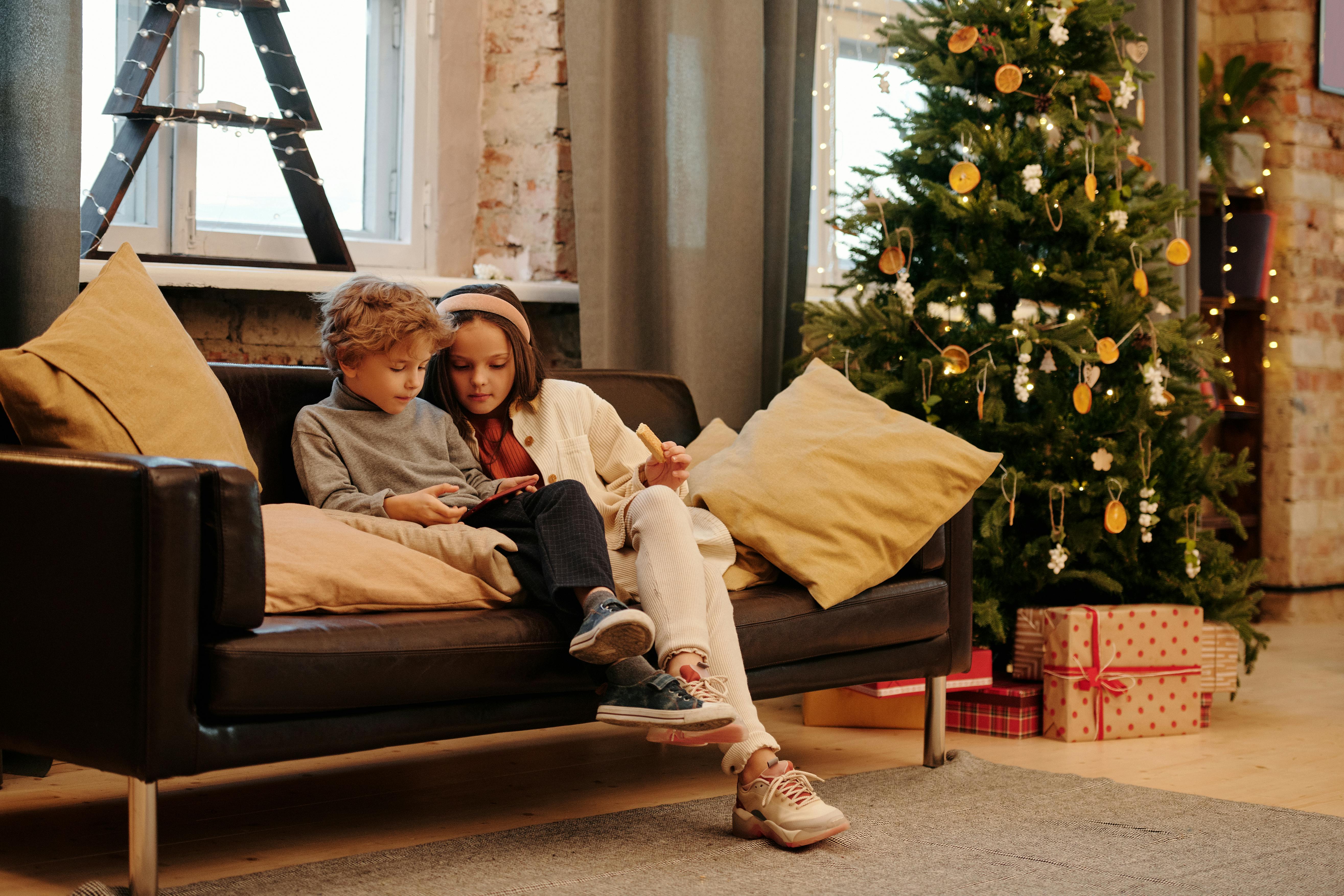 Zwei Kinder sitzen auf einer Couch in einem weihnachtlich dekorierten Zimmer und schauen zusammen in ein Buch.