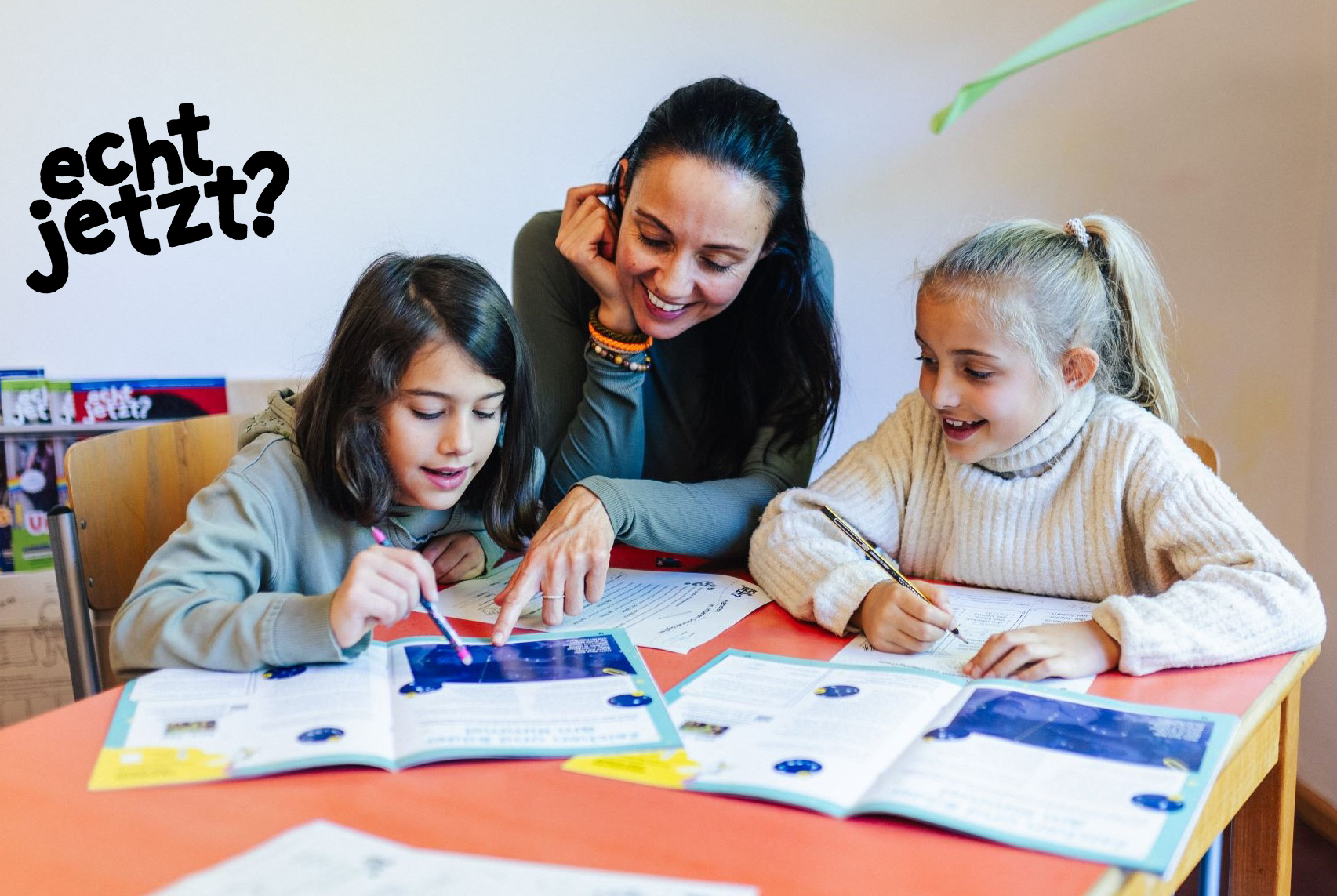Lehrkraft sitzt mit zwei Kindern am Tisch und schaut in das echt jetzt Magazin